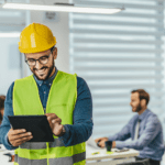 A man wearing a yellow hard hat is looking down at an iPad smiling.